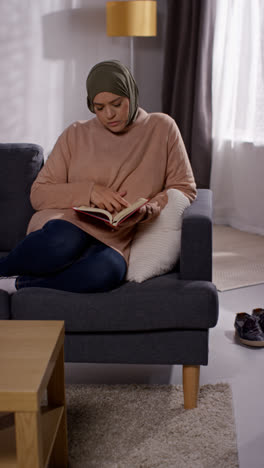 Vertikales-Video-Einer-Muslimischen-Frau-Mit-Hijab,-Die-Zu-Hause-Auf-Dem-Sofa-Sitzt-Und-Den-Koran-Liest-Oder-Studiert